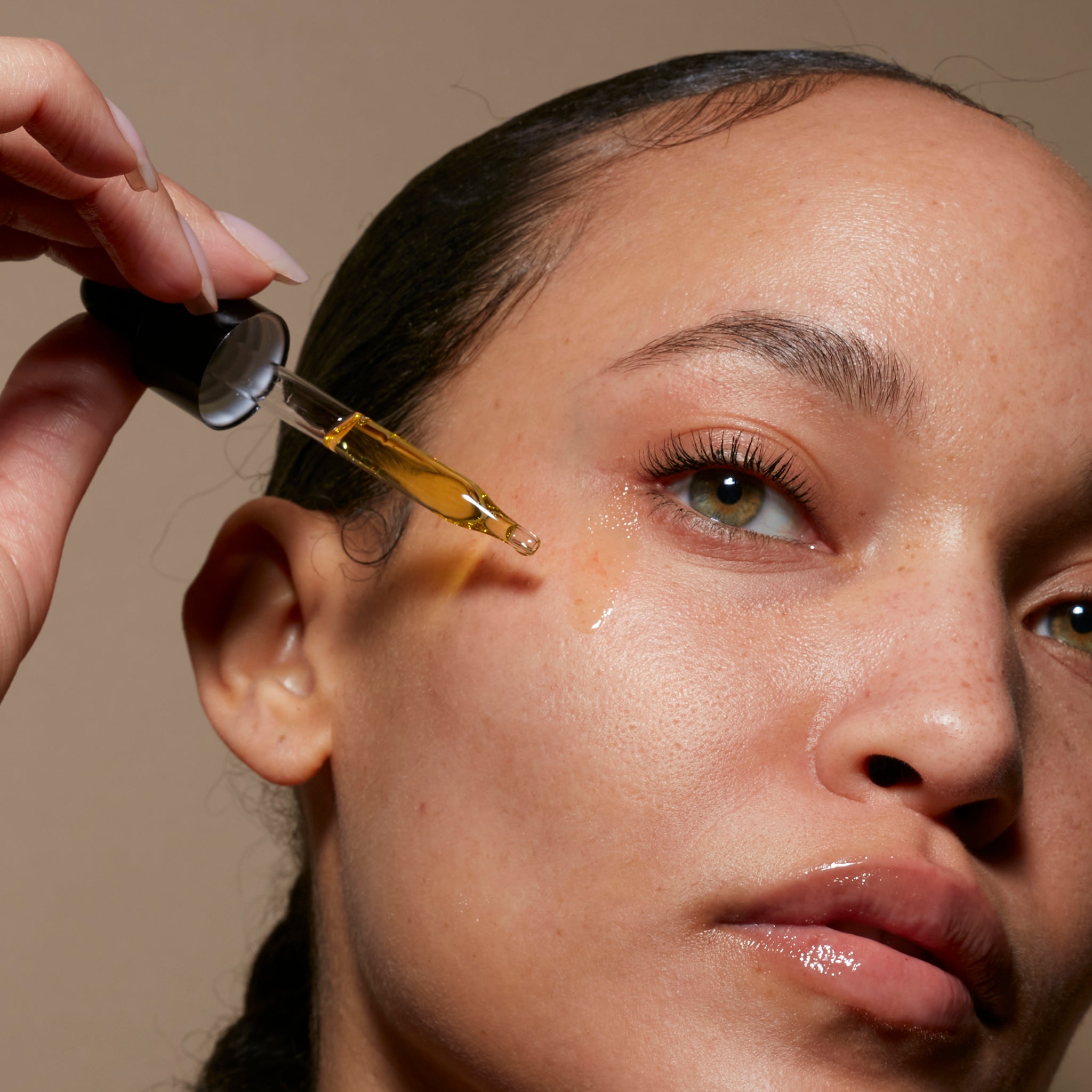 Close-up of a woman using the eyedropper to apply Le Prunier Plum Beauty Oil on her cheek, with neutral lighting.