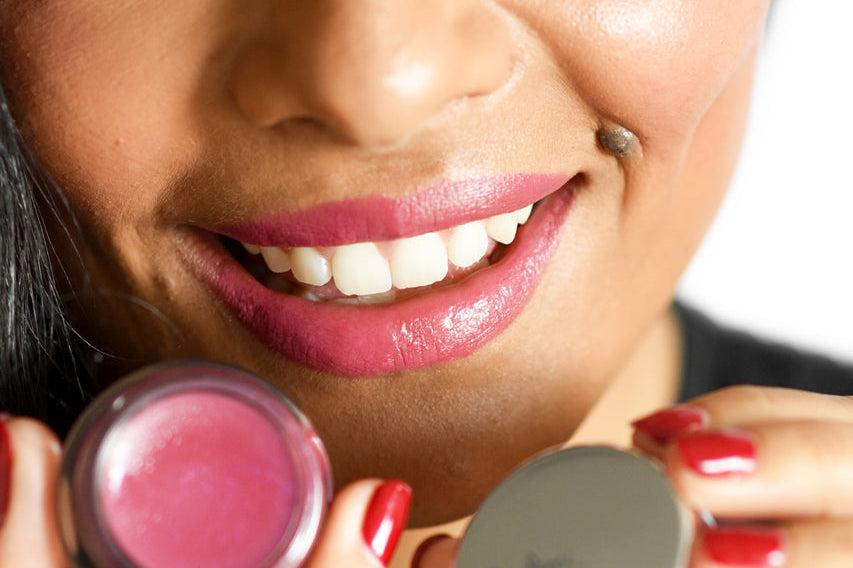 A smiling woman with medium complexion and golden undertones holding the Ere Perez Holy, which she applied to her lips.