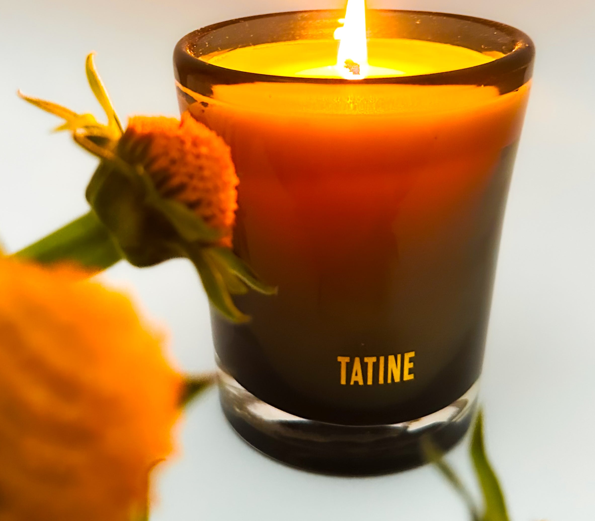 A lit Tatine candle in its glass tumbler shedding light on a bed of flowers.