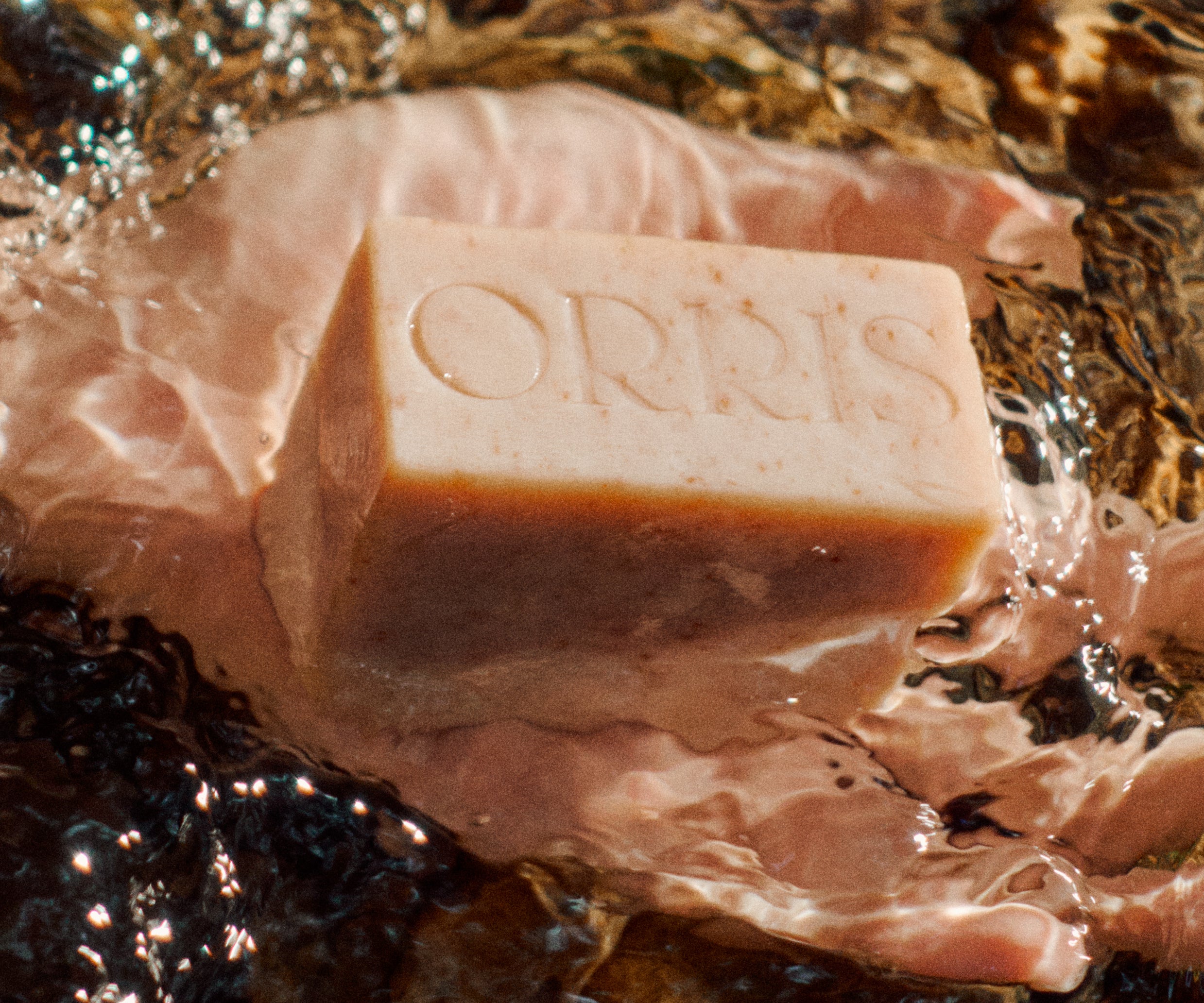 A hand holding a light tan ORRIS Cleansing Bar partially submerged in clear, rocky stream water.