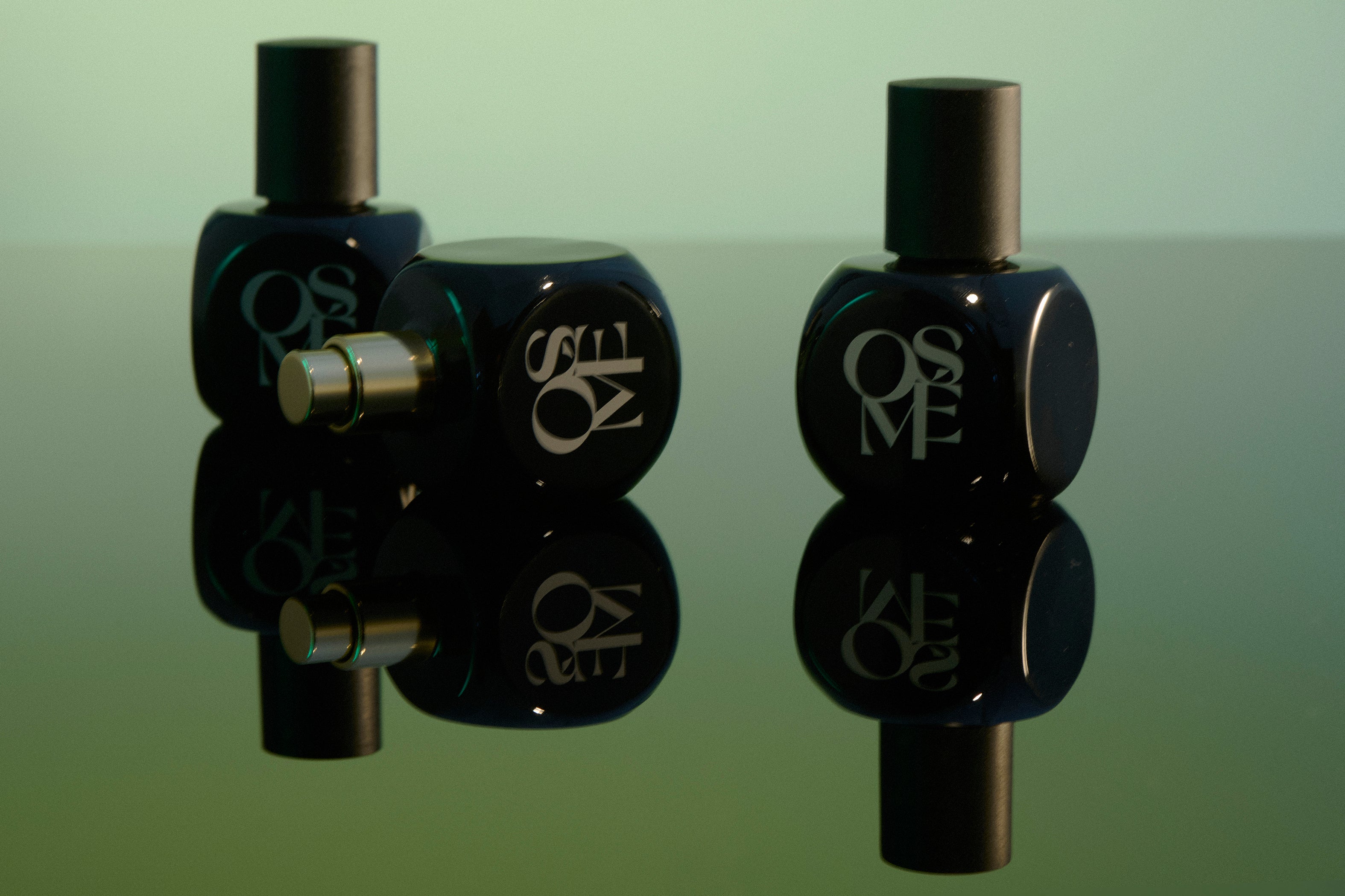 
  
  Osmé Fragrances displayed black perfume bottles with caps on and off with a green back ground and mirrored counter showing reflection.
  
