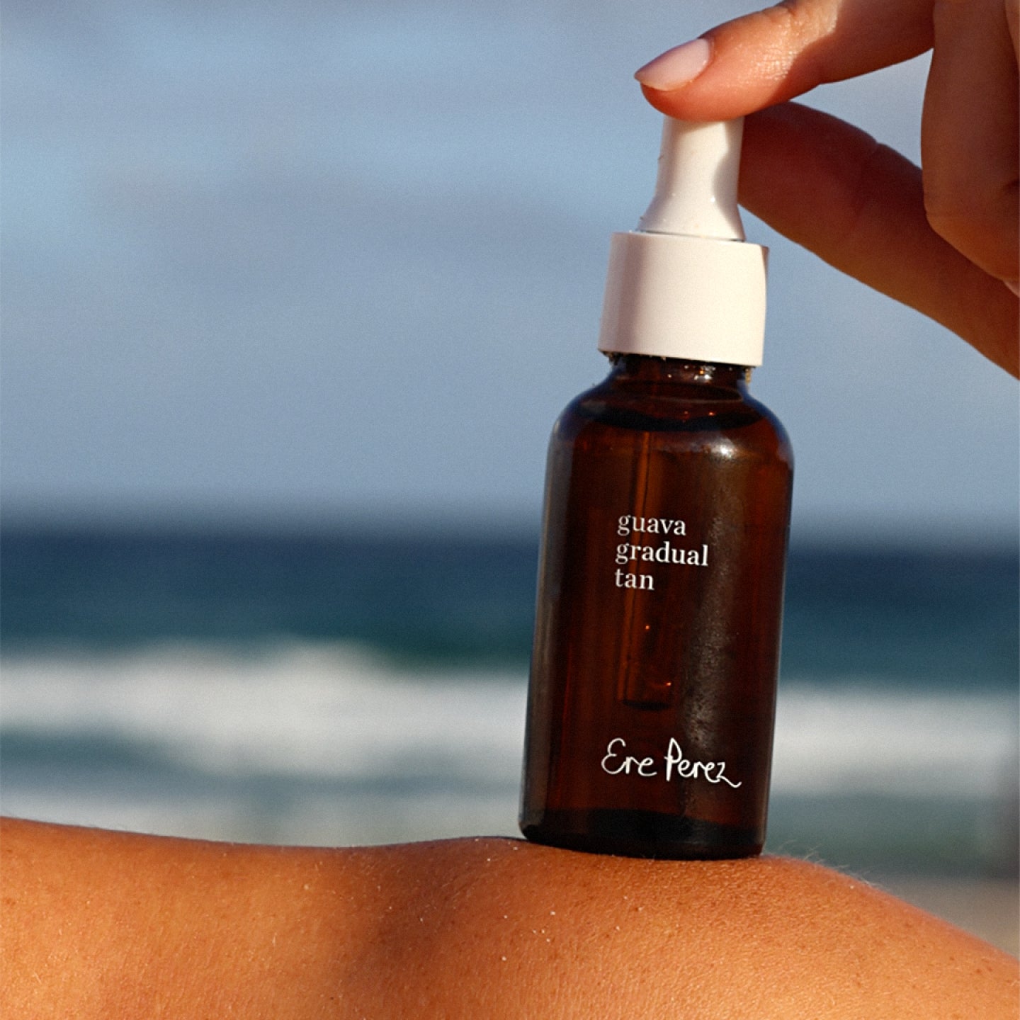 
  
  Brown bottle labeled 'guava gradual tan' by Ere Perez held over skin with ocean background
  
