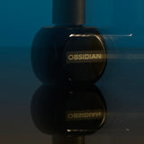 
  
  Black Osmé Fragrance Obsidian perfume bottle on a reflective surface with a blue background
  
