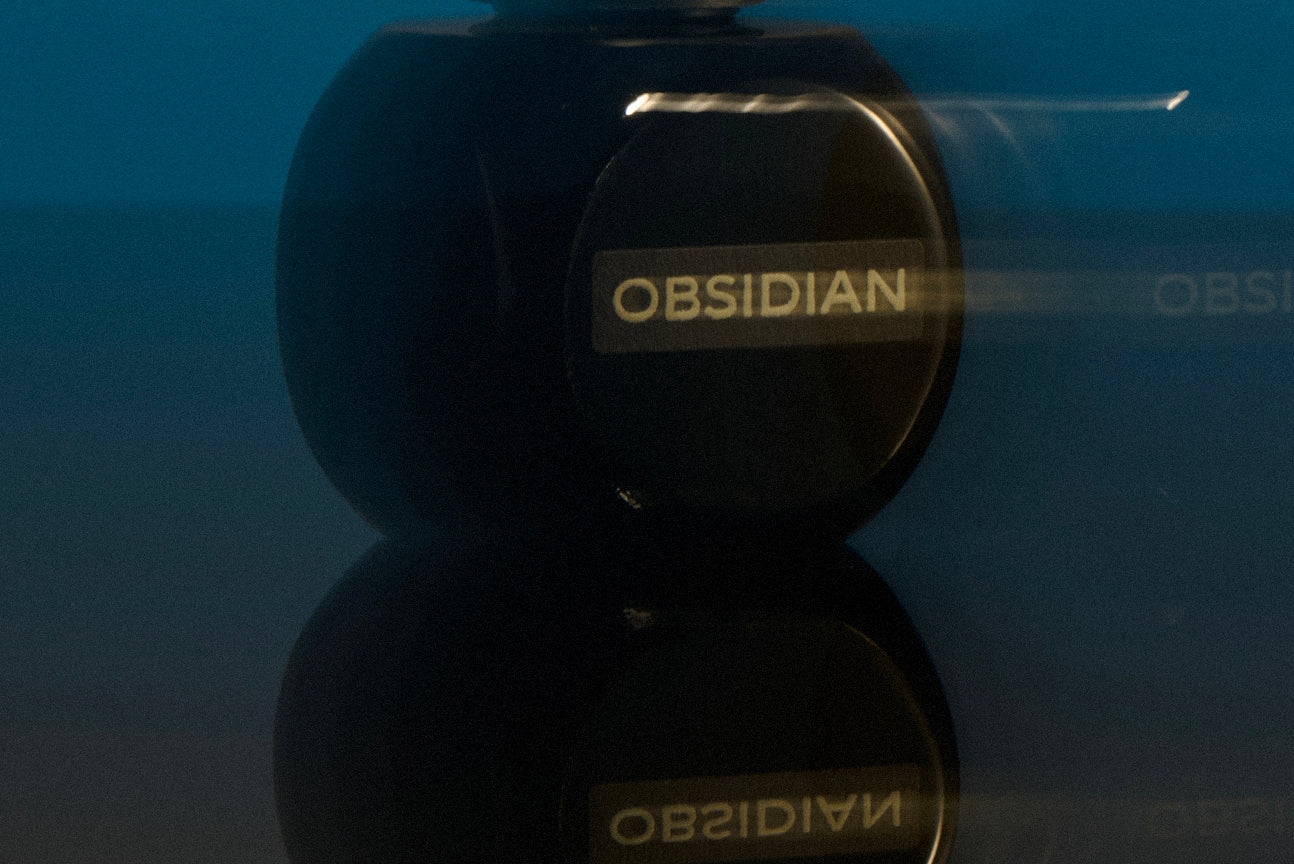 
  
  Black Osmé Fragrance Obsidian perfume bottle on a reflective surface with a blue background
  
