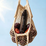 
  
  Net bag with plums and a bottle of Plum Spritz against a clear blue sky.
  
