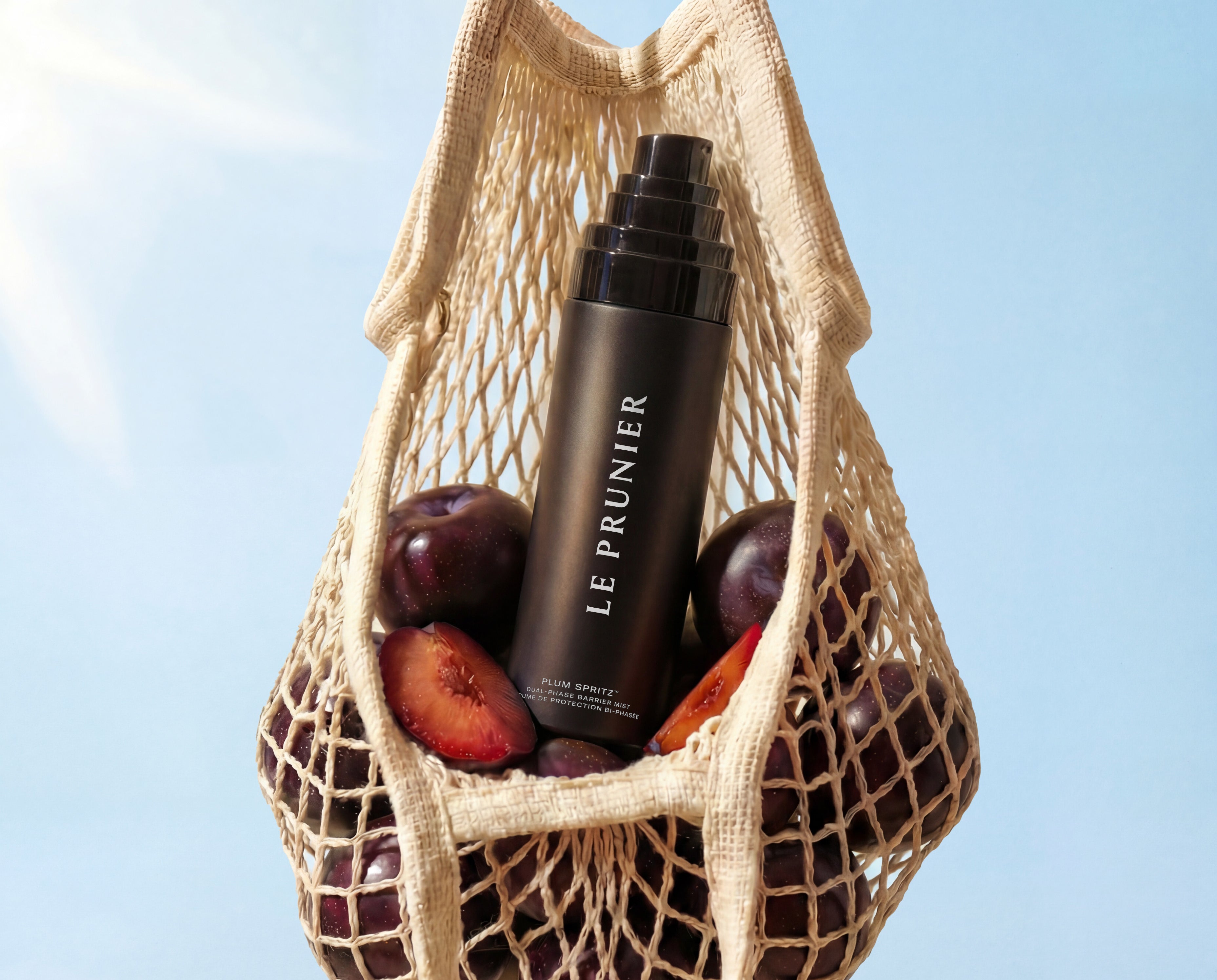 
  
  Net bag with plums and a bottle of Plum Spritz against a clear blue sky.
  
